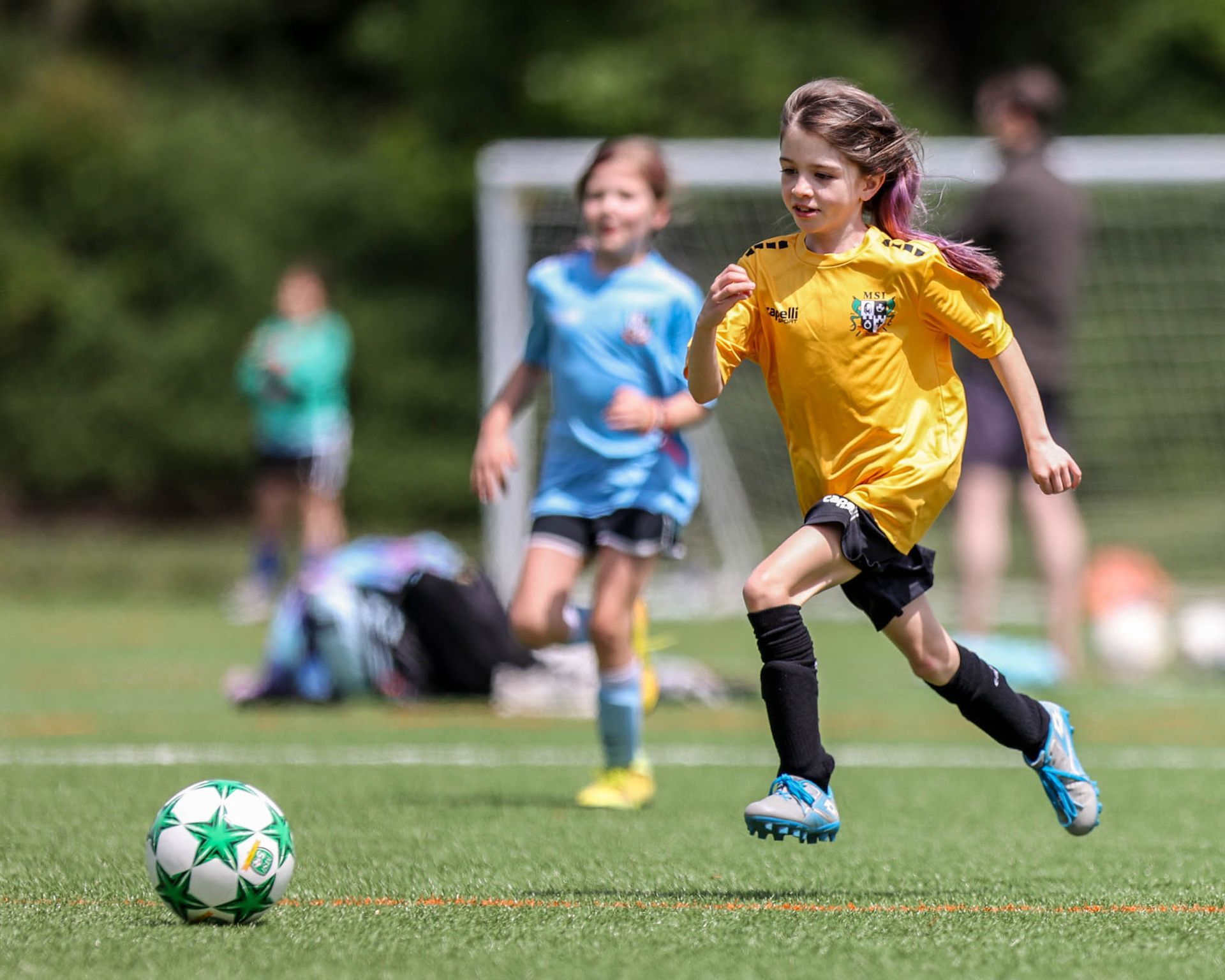 Girl in yellow MSI soccer uniform sprints toward ball during youth match as opponent in blue runs close behind on turf field.