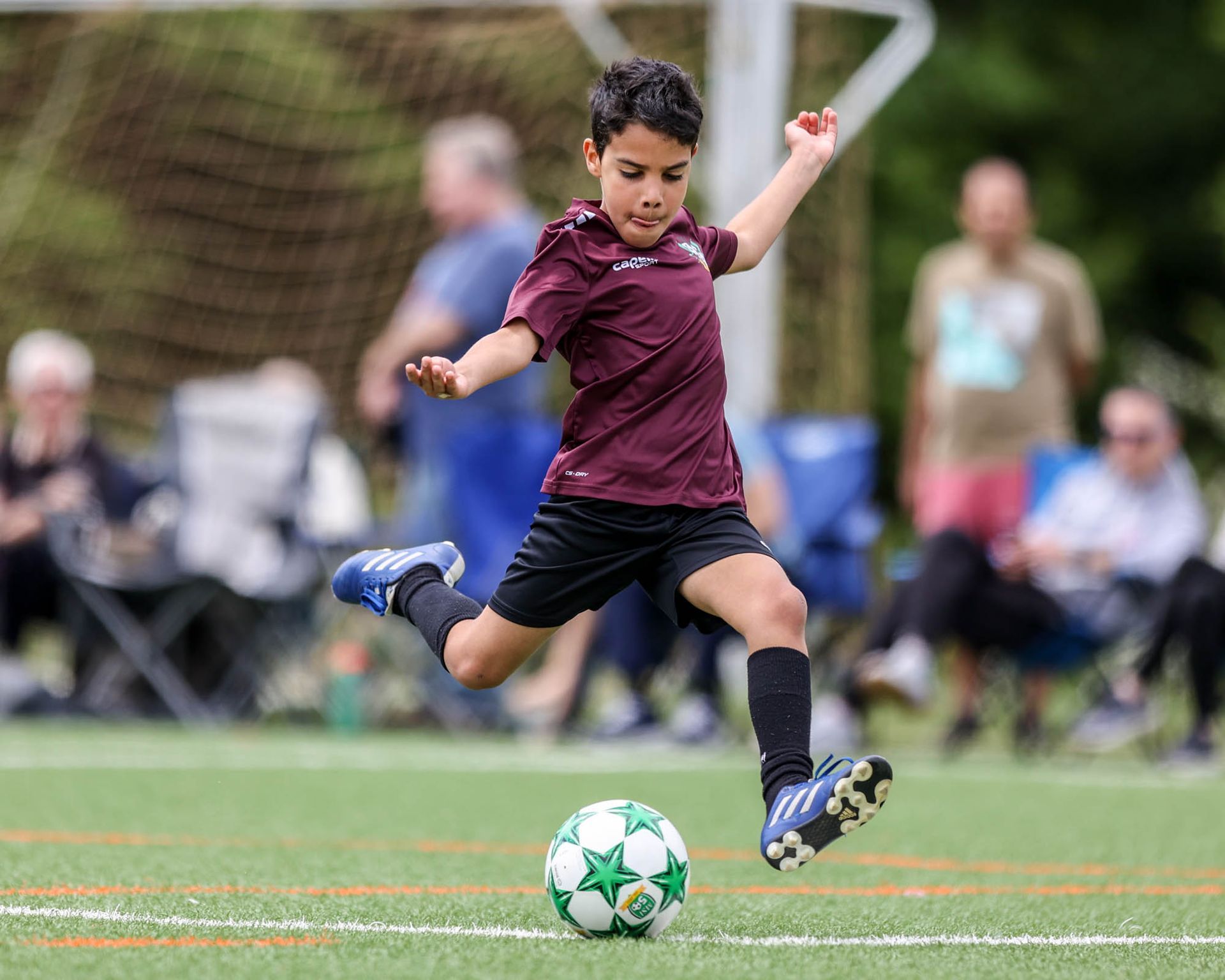 Youth MSI soccer player in maroon uniform prepares a strong kick on turf field as spectators sit and watch the match.