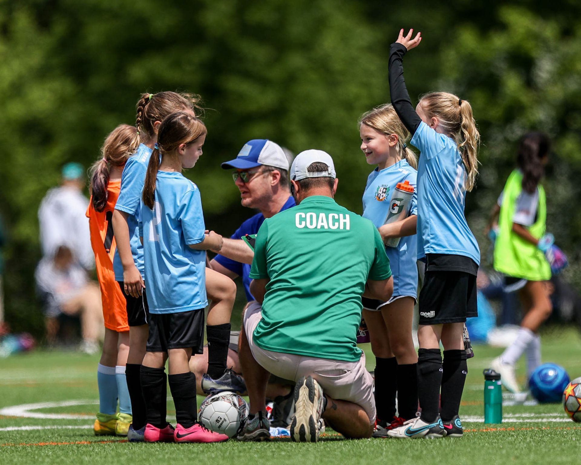 MSI soccer coach kneels with young girls in blue uniforms on turf field, giving instructions during youth practice.