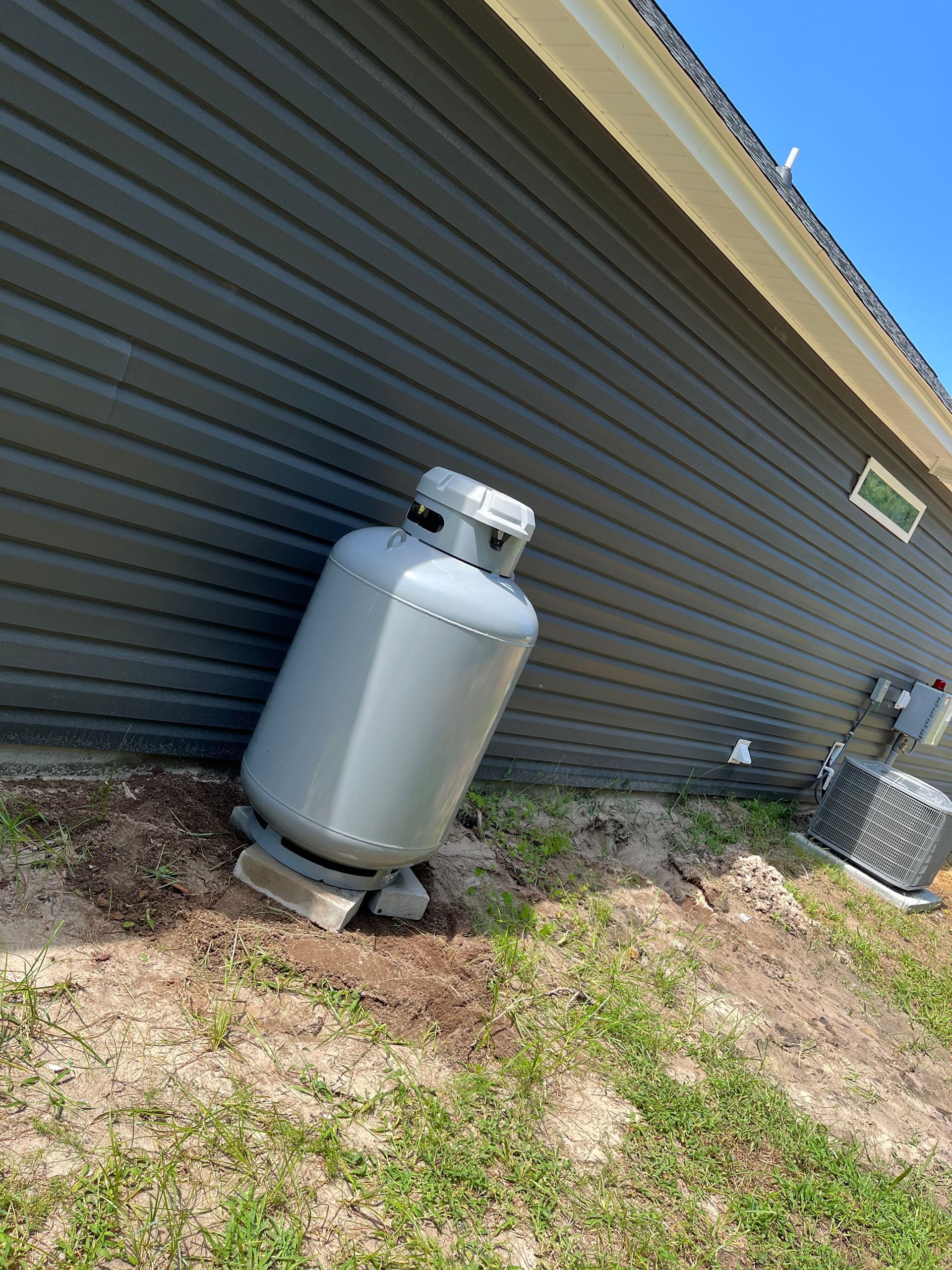 A tall, light-gray propane tank sits on a concrete block next to the gray-sided wall of a house near an AC unit.