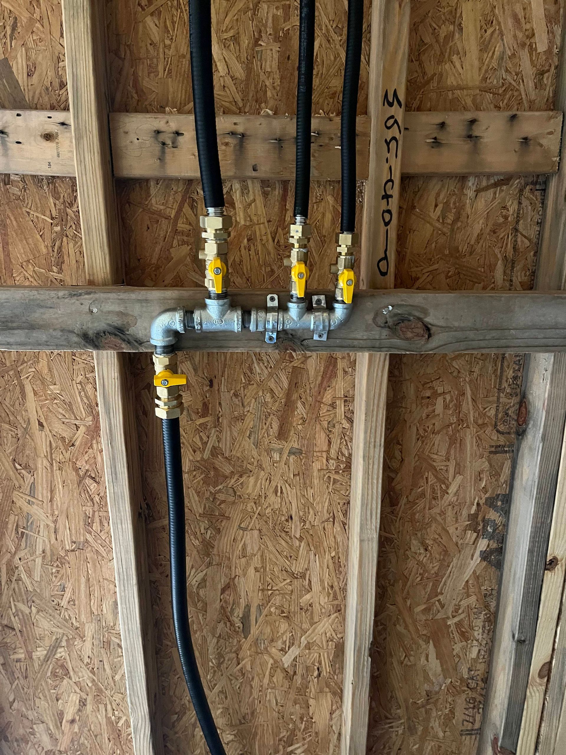 A gas manifold system with four yellow-handled shut-off valves and black flexible piping mounted to wooden wall studs.