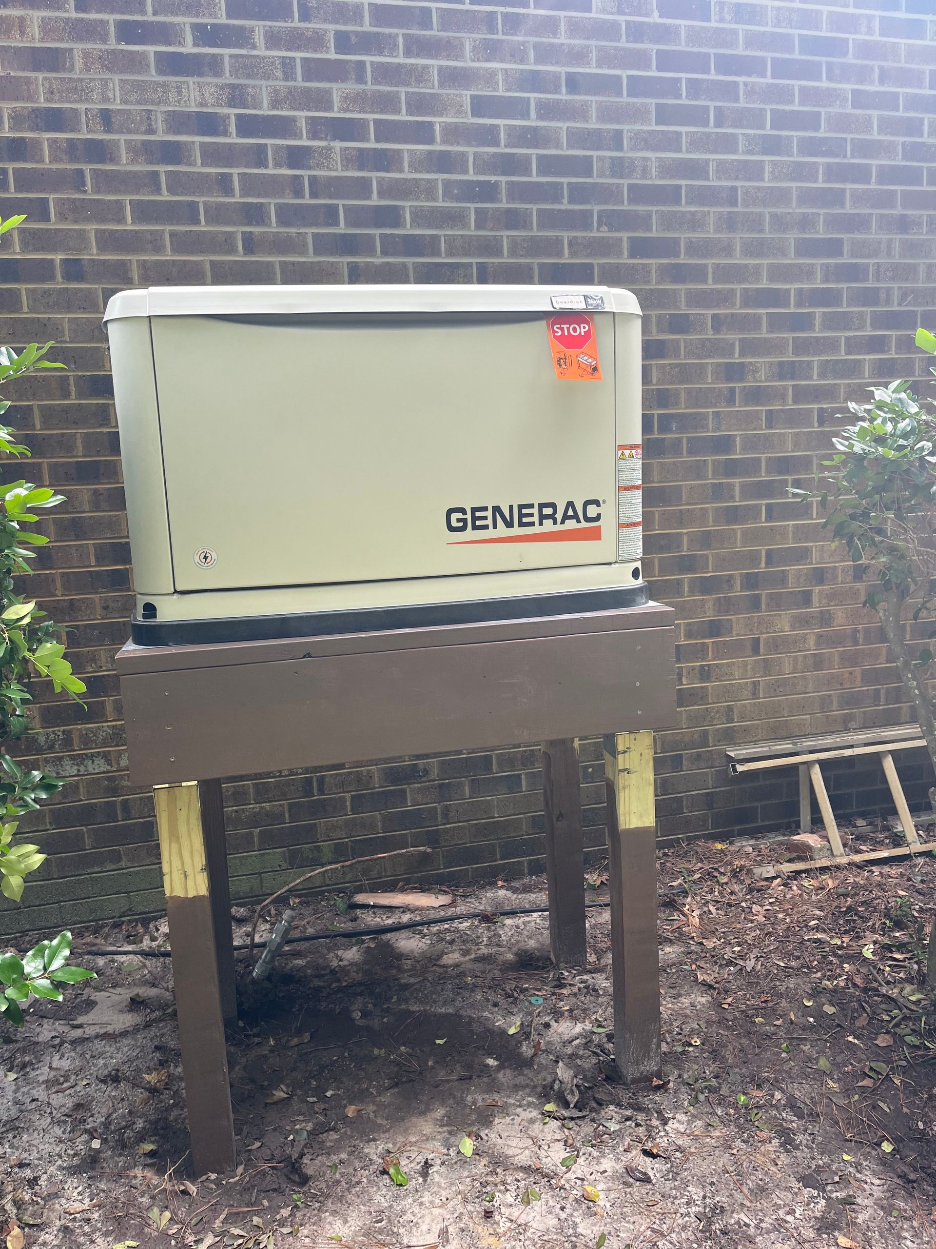 A beige Generac standby generator mounted on a raised wooden platform against a brick wall.