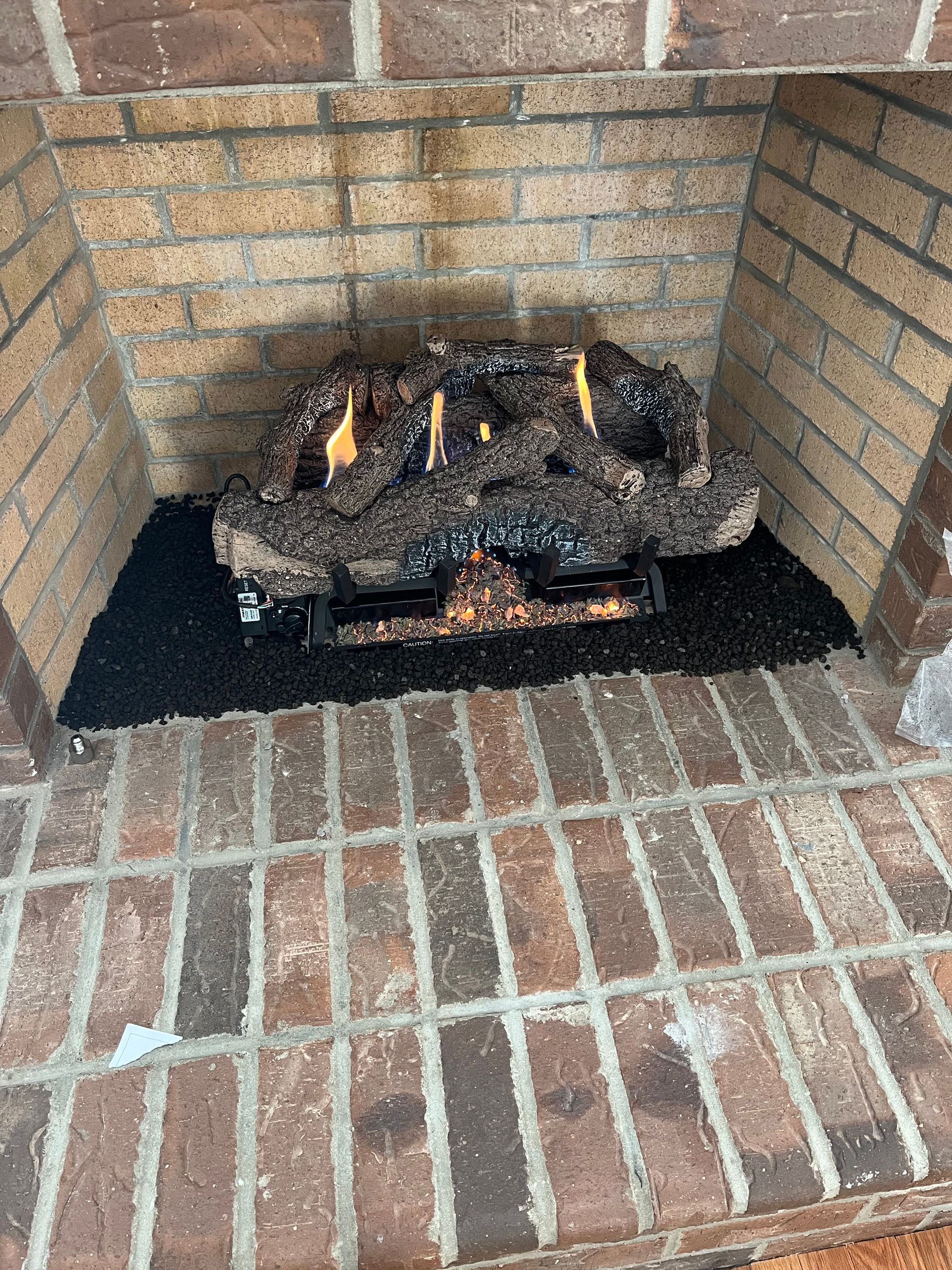 A gas fireplace insert with burning logs sits inside a brick fireplace hearth covered with black fire glass.