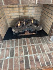 A gas fireplace insert with burning logs sits inside a brick fireplace surround on a base of black fire glass.