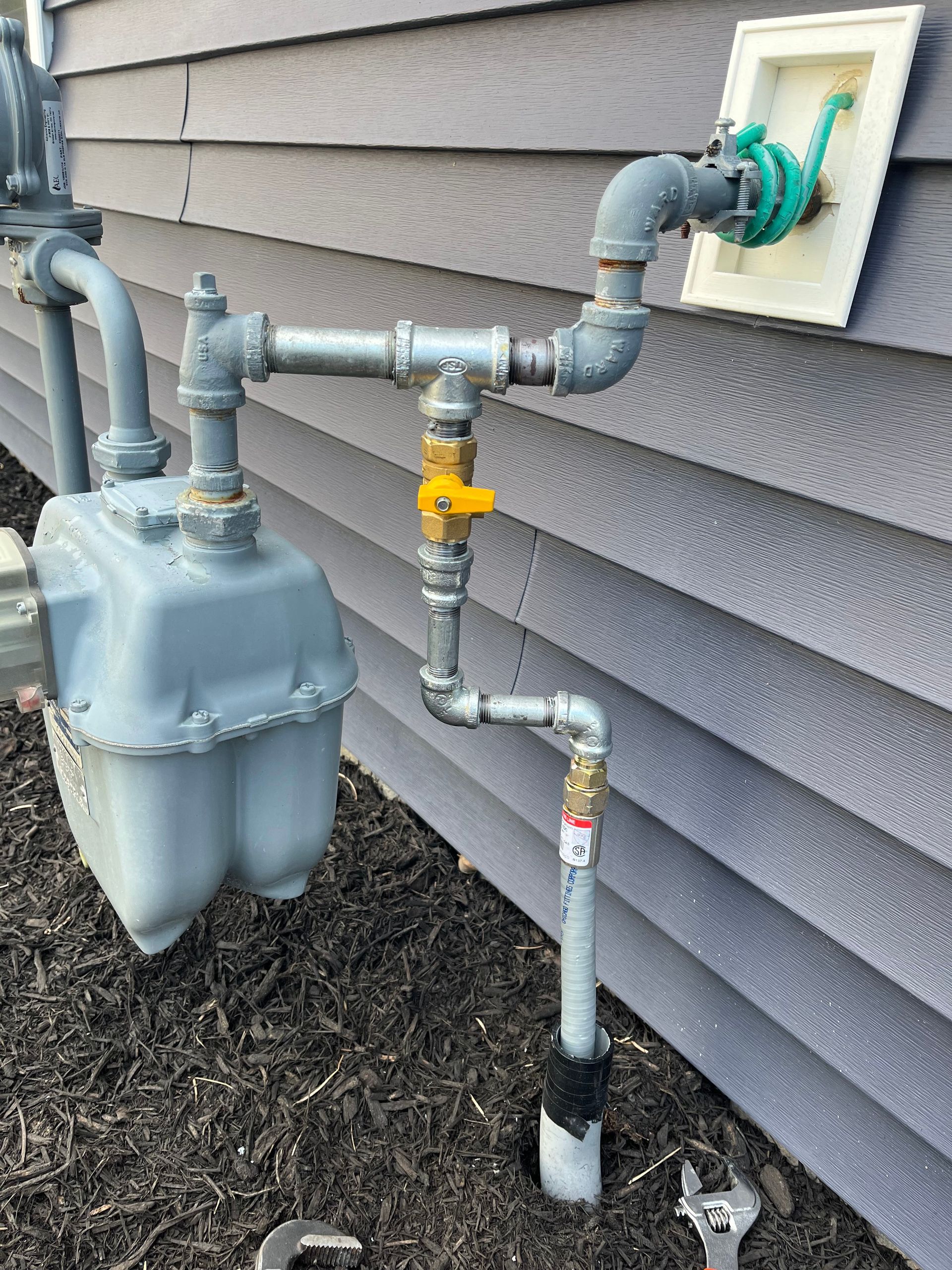 A gray gas meter and metal piping installed against dark gray siding, including a yellow-handled shut-off valve.