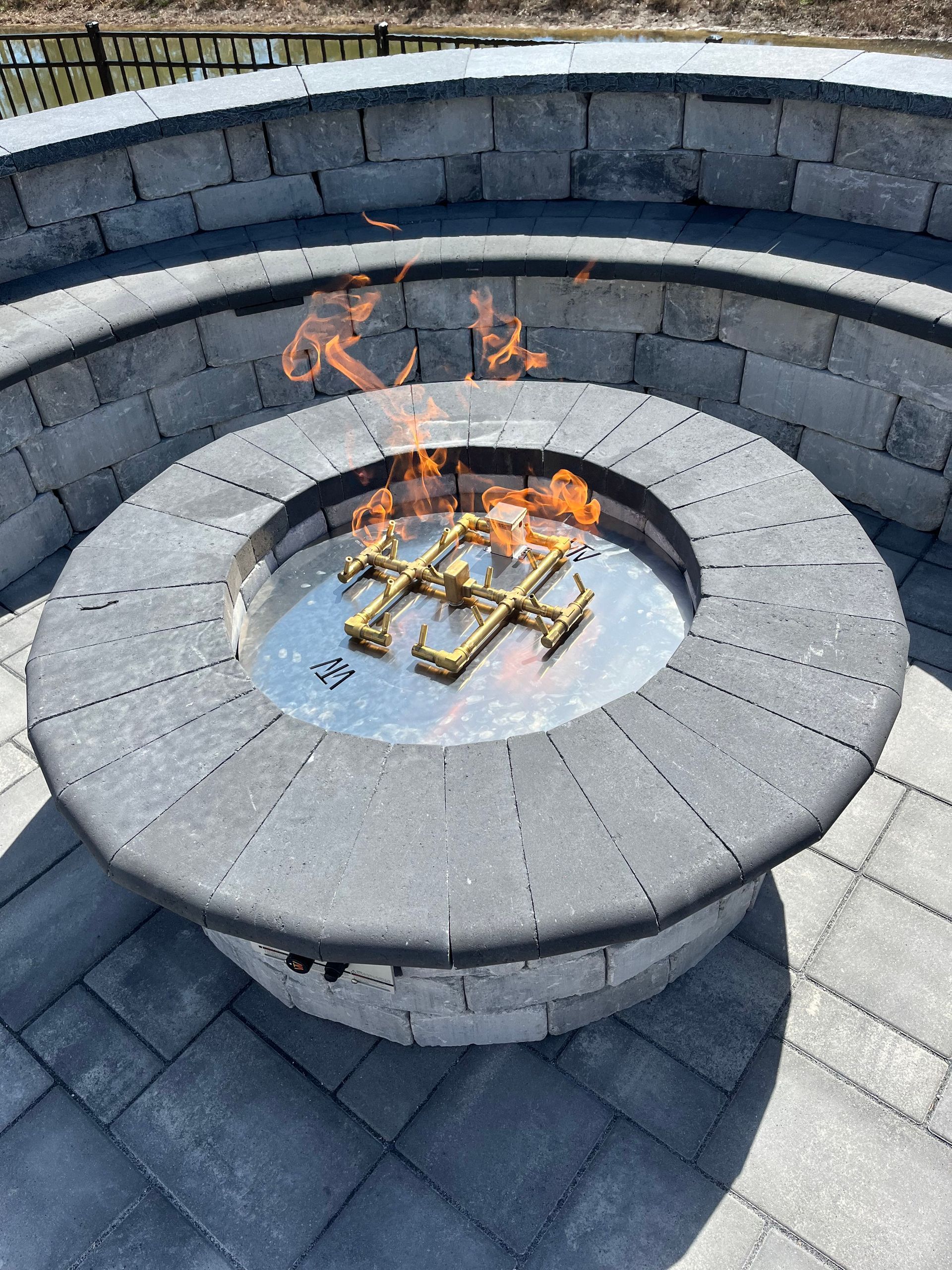 A lit circular stone fire pit centered on a matching stone patio with a curved stone seating wall in the background.