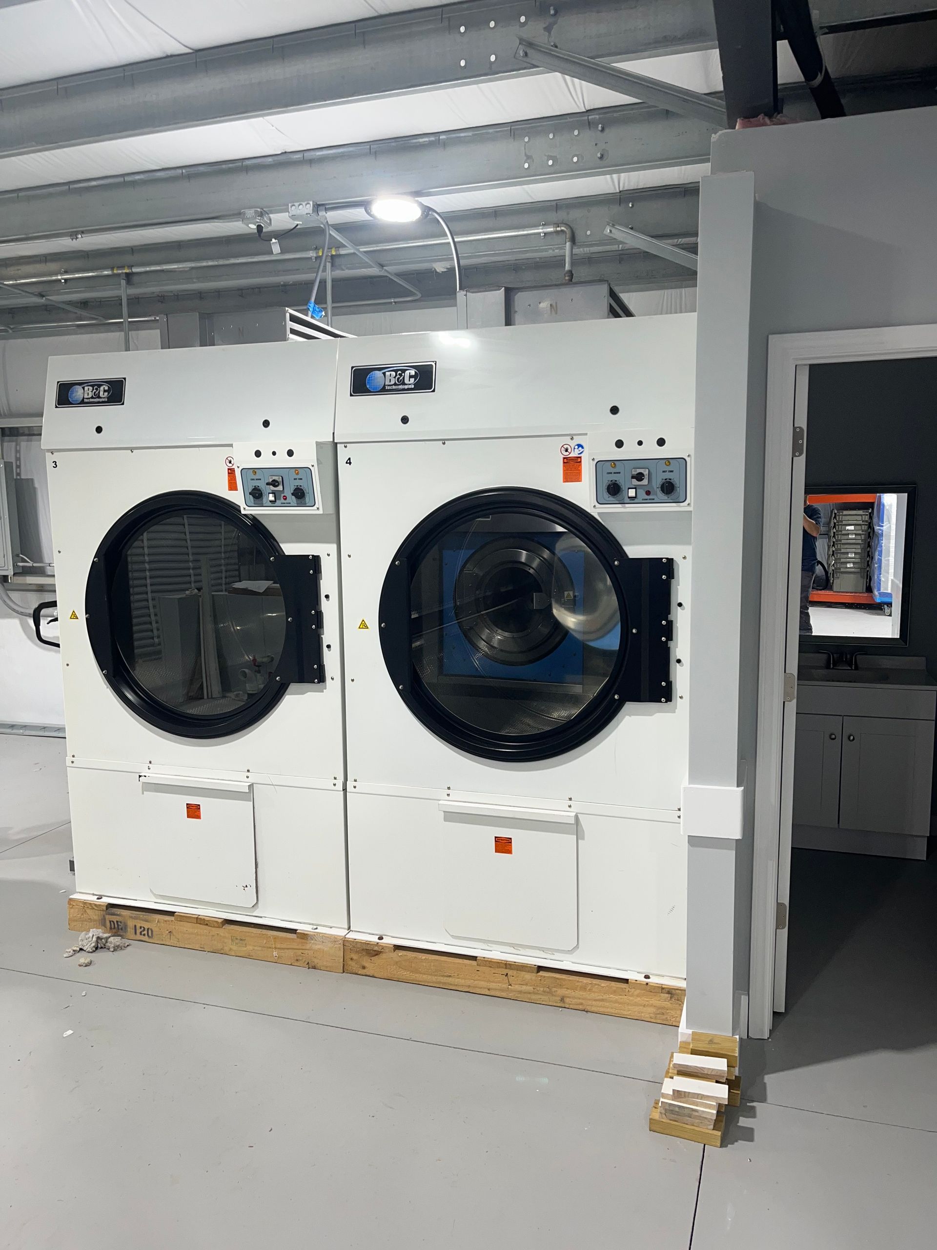 Two large, white industrial tumble dryers sit side-by-side in a commercial laundry facility with grey flooring.