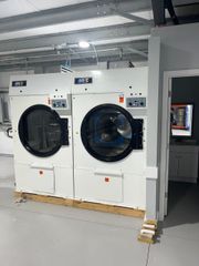 Two industrial white laundry dryers sit side-by-side in a brightly lit, open-plan facility.