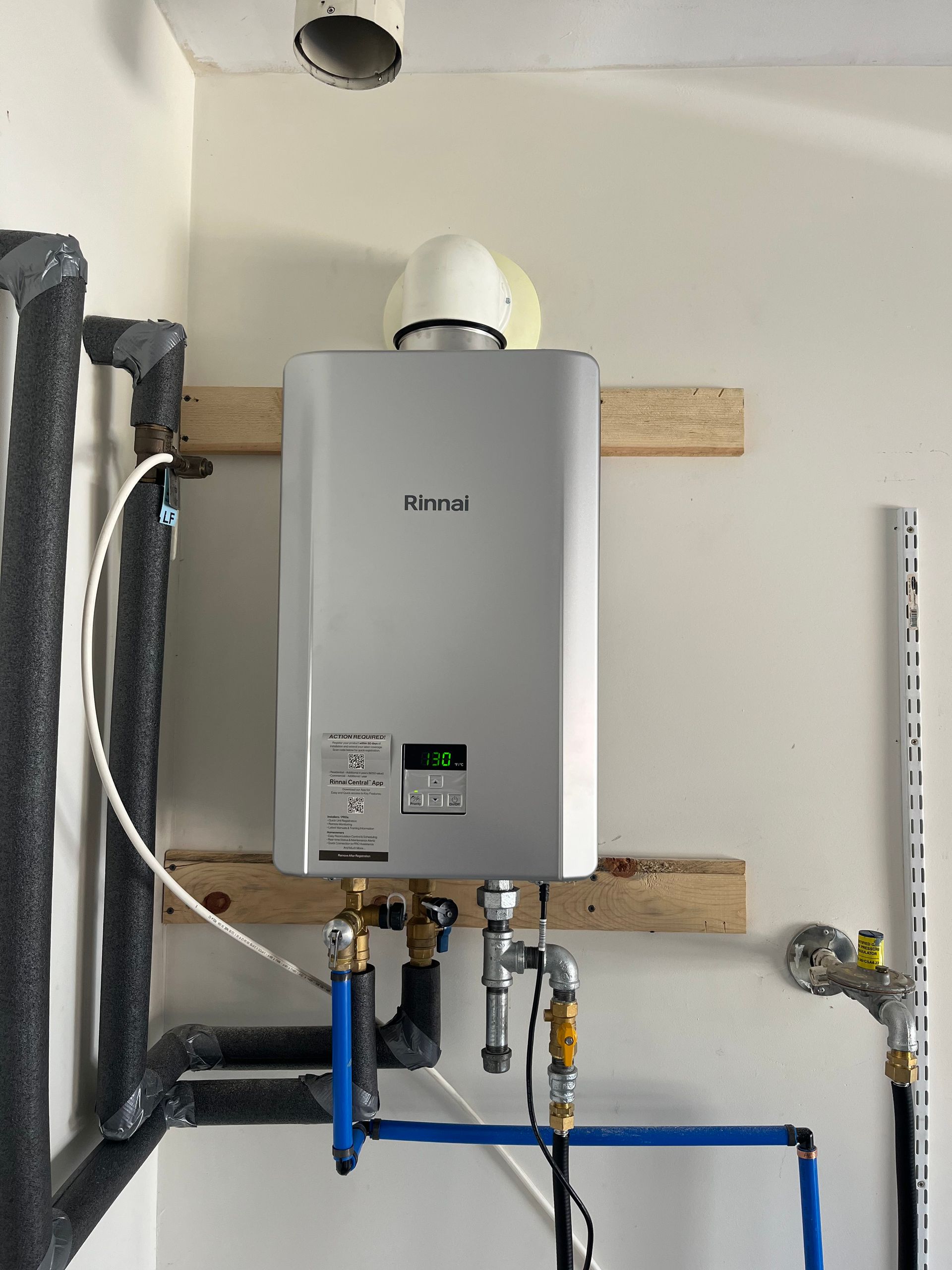 A silver Rinnai tankless water heater mounted on a wall with plumbing pipes below and a vent pipe above.