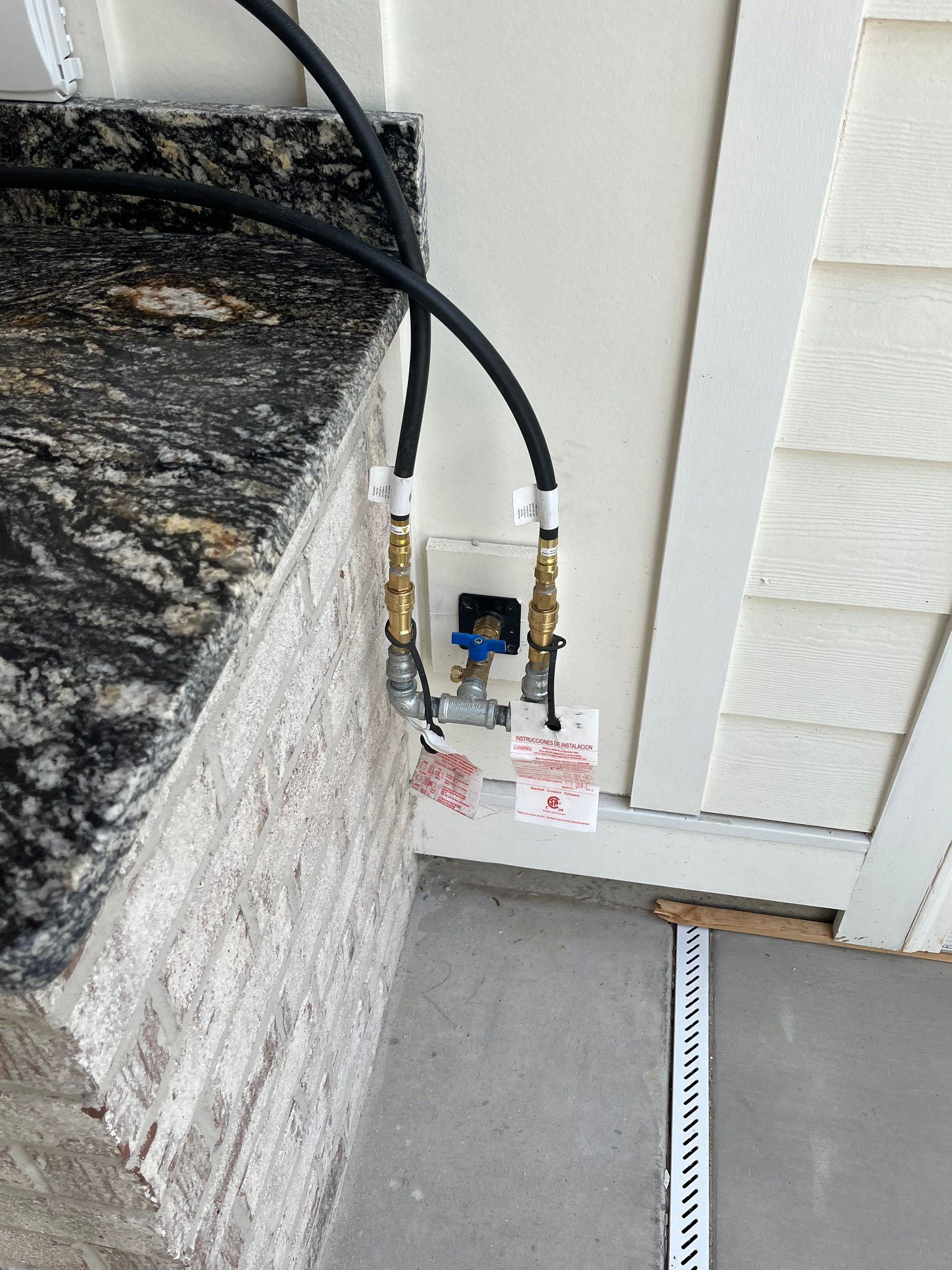 Two black gas hoses connected to brass valves on an outdoor wall next to a stone counter and white siding.
