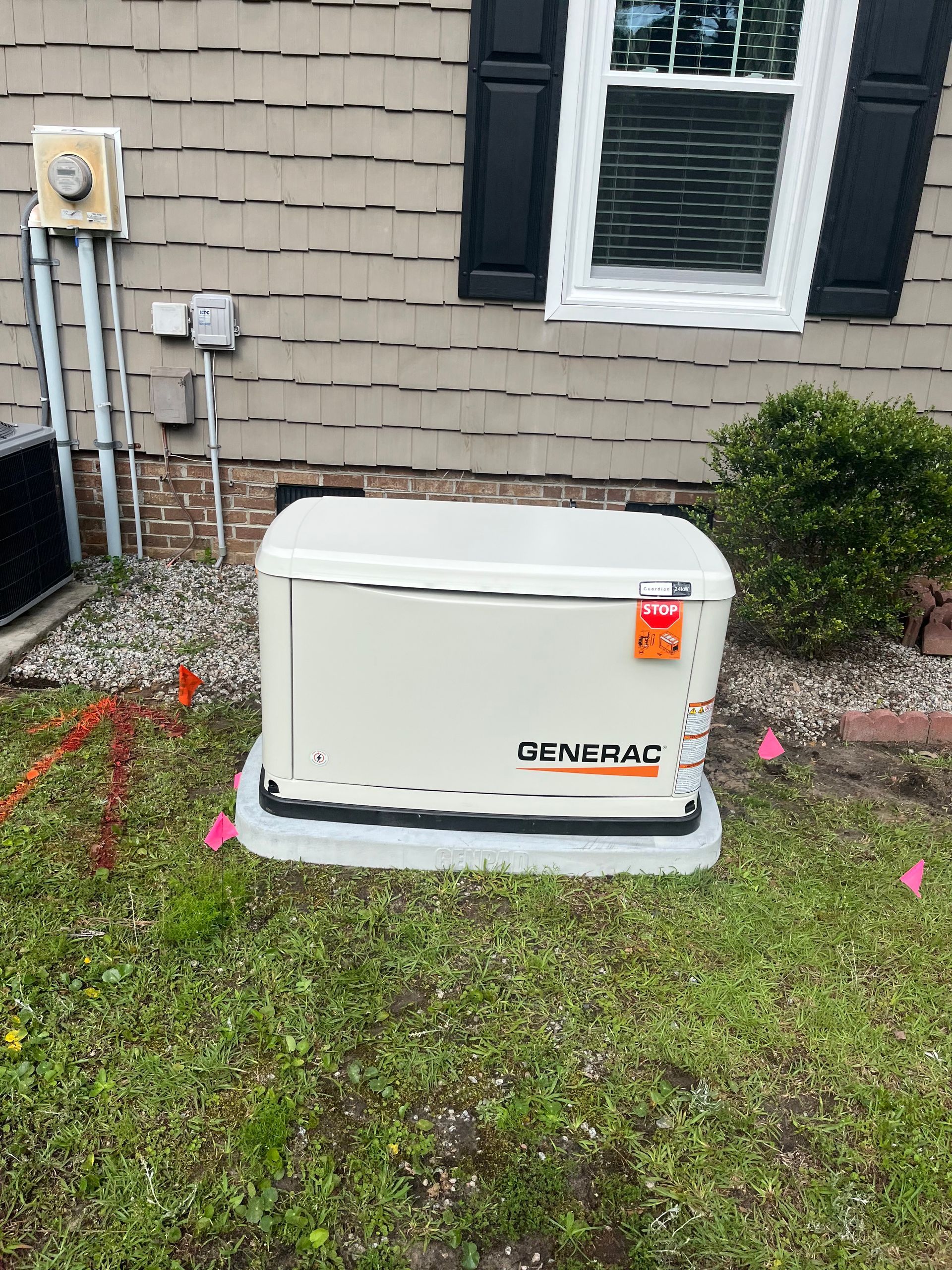 A beige Generac backup generator sits on a concrete pad outside a house next to a window and utility conduits.