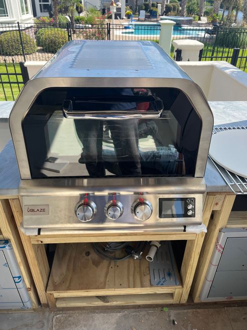 A stainless steel outdoor grill with a glass-front hood, installed in a wooden framing outdoors near a swimming pool.