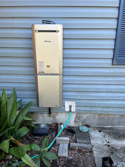 A gold-colored tankless water heater mounted on a house exterior siding above a garden hose connected to a wall spigot.