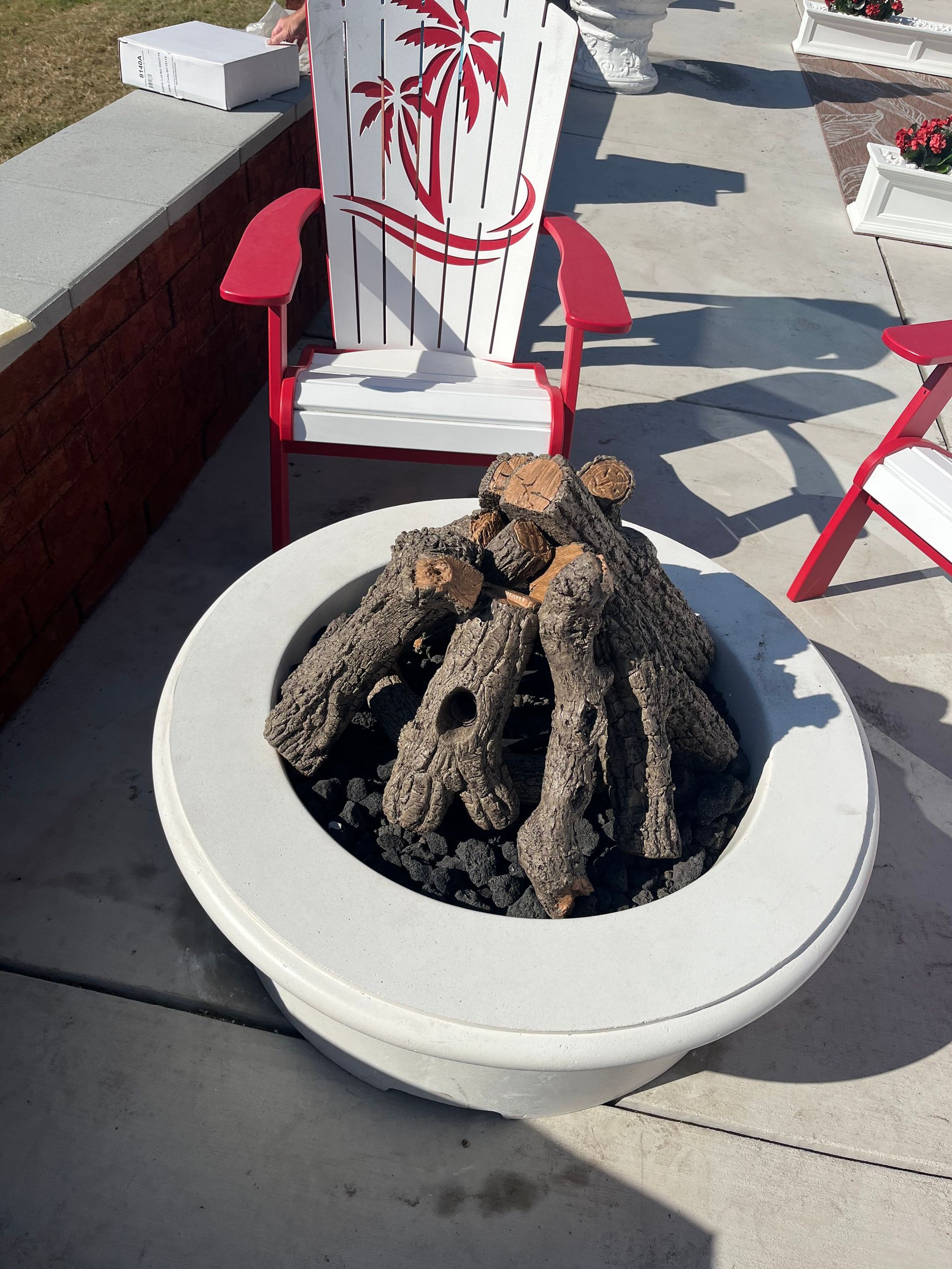 A circular concrete fire pit with artificial logs sits on a patio next to an Adirondack chair featuring a palm tree design.