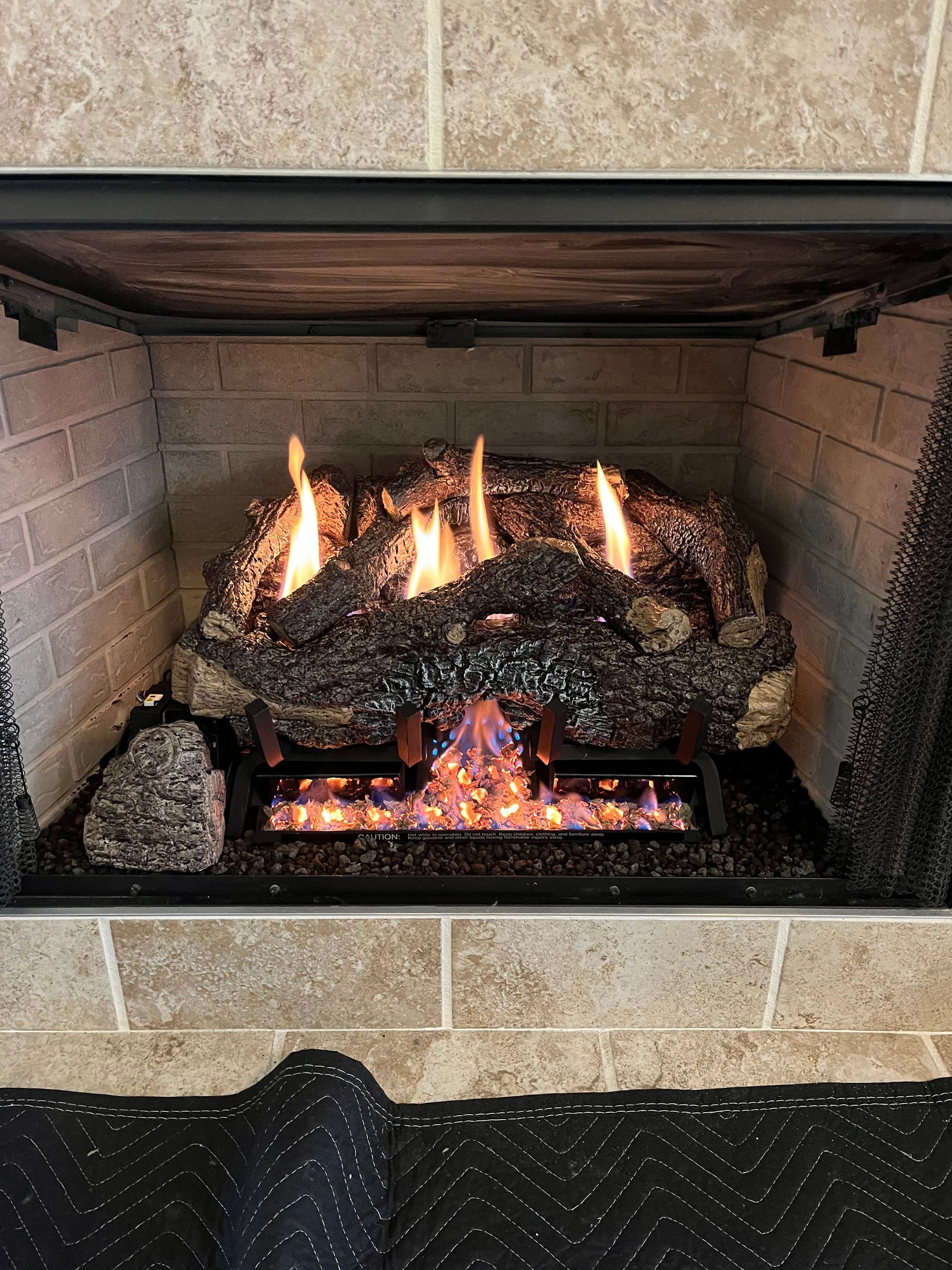 A gas fireplace insert with burning logs and glowing embers inside a stone-tiled hearth.