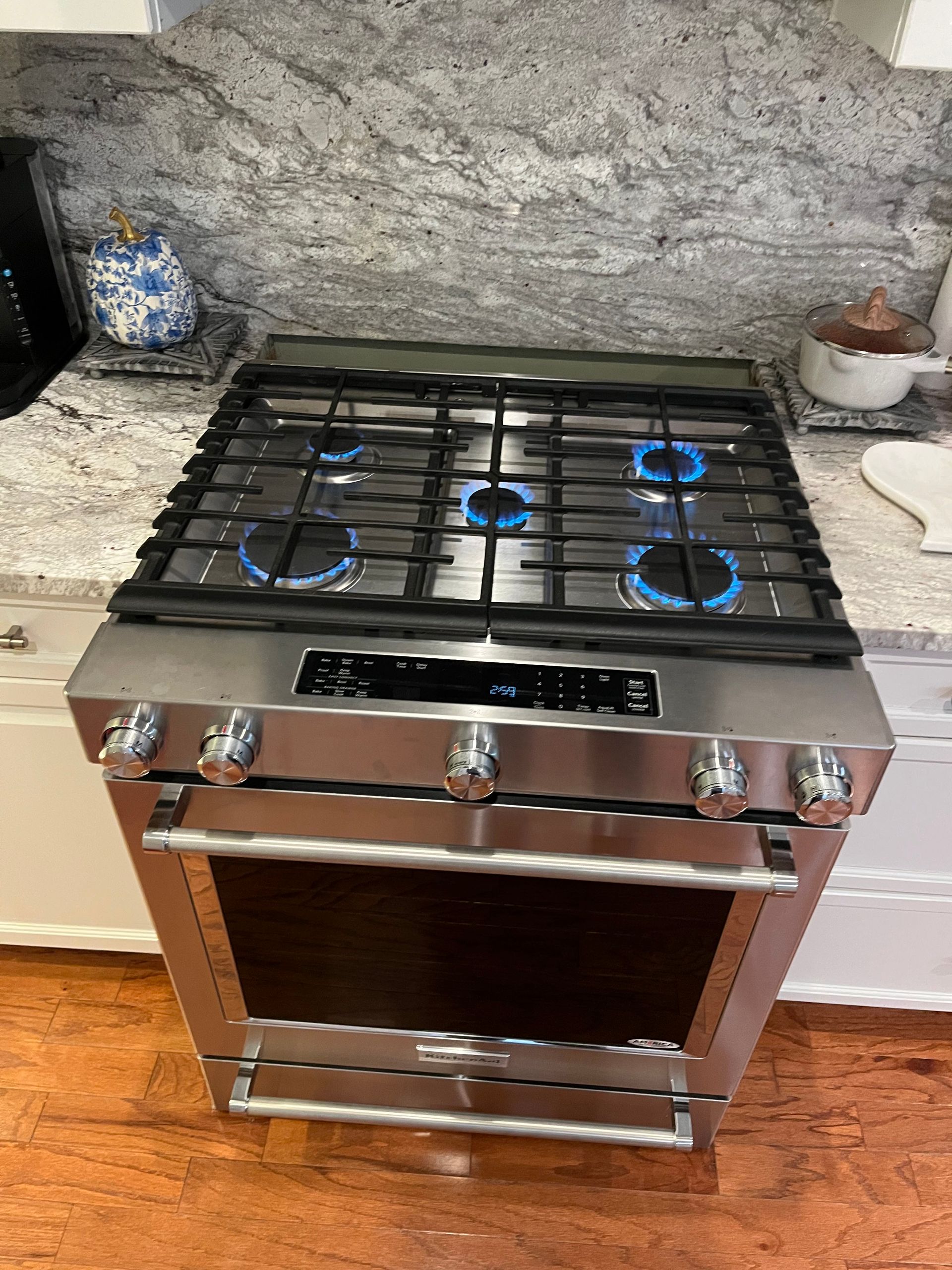 A stainless steel gas range with four active blue flames, situated in a kitchen with granite countertops.