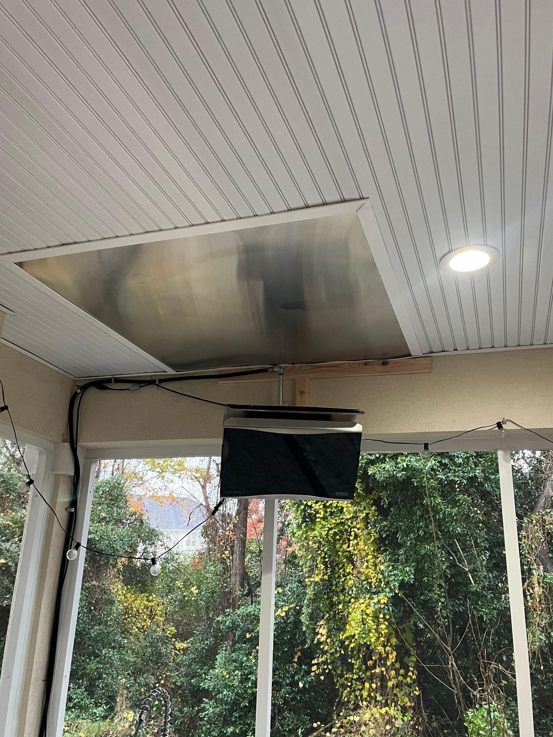 A porch ceiling featuring a rectangular metal panel insert, a recessed light, and a hanging projector under a roof.