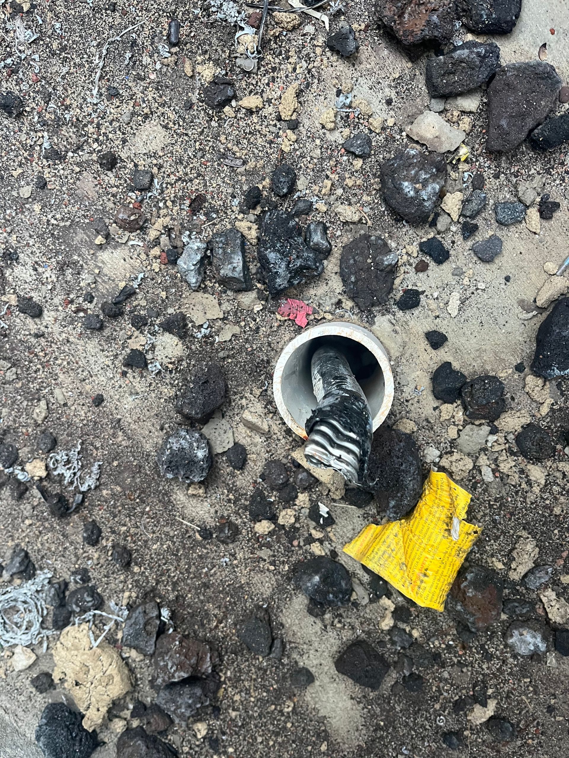A white plastic pipe sticking out of dirt and gravel, with dark, layered material inside and a yellow scrap nearby.