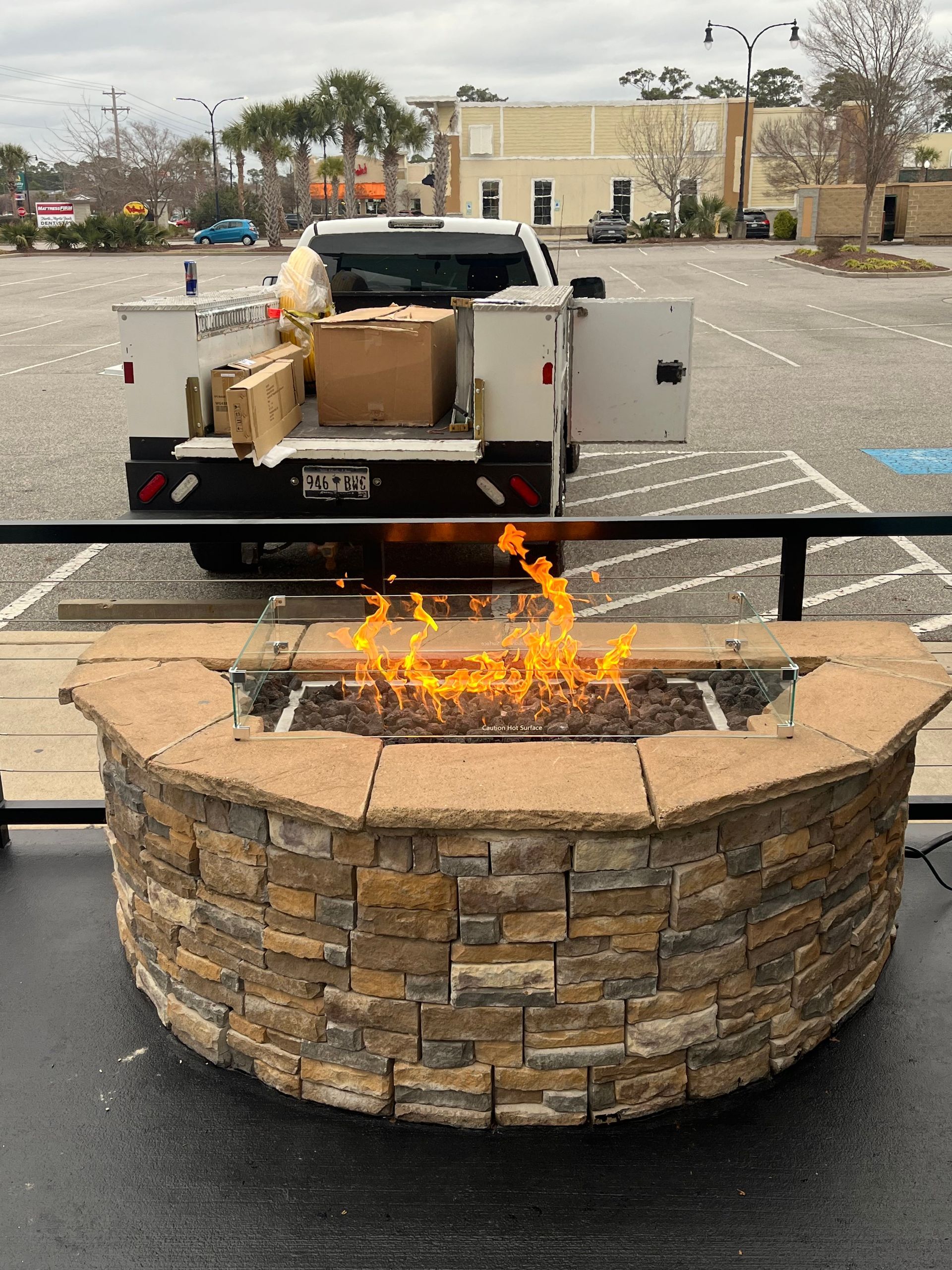 A stone fire pit with flickering orange flames outdoors, with a parked utility truck visible in the background.