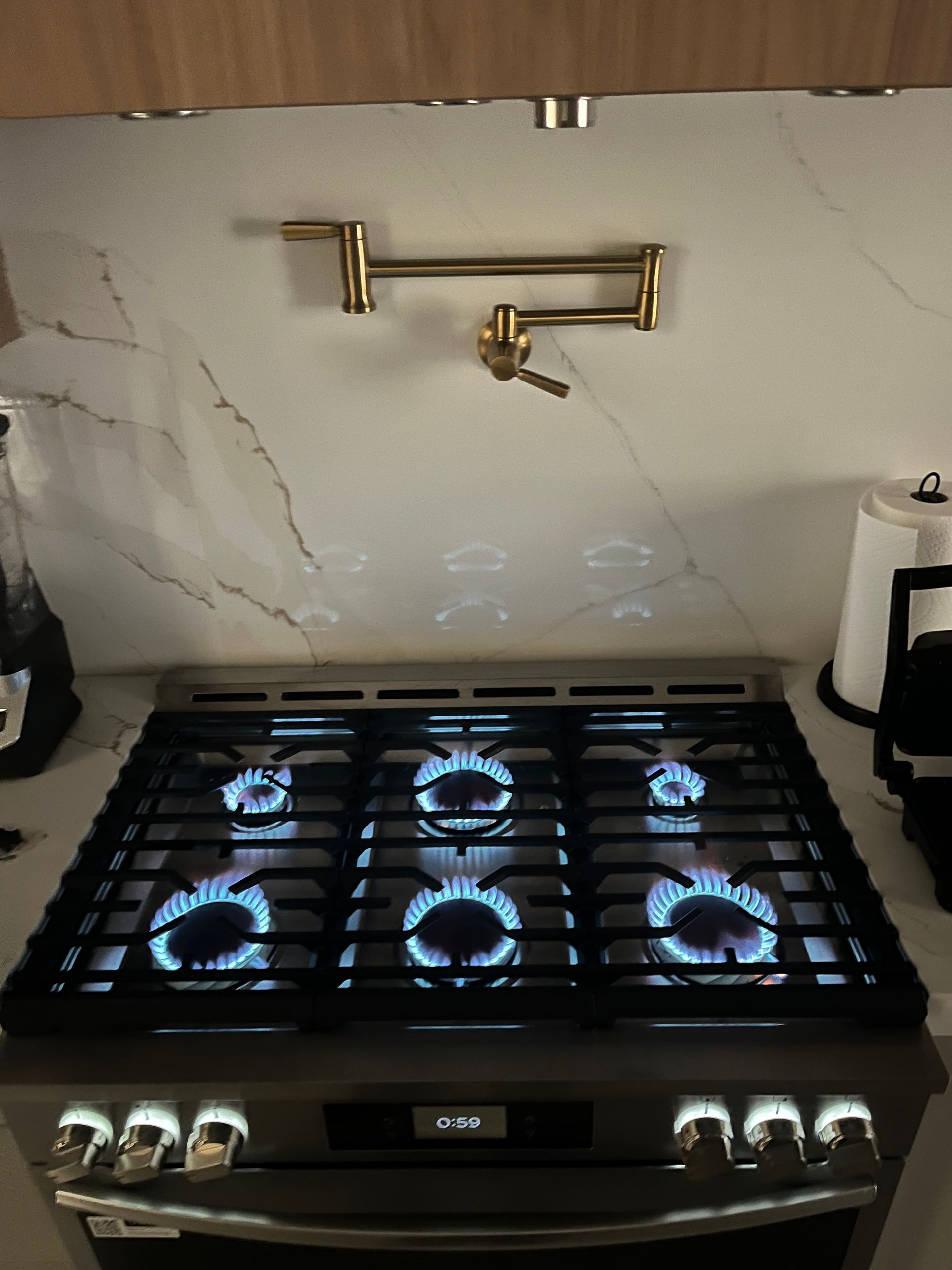 A brass pot filler faucet is mounted on a white marble-patterned backsplash above a gas stove with all burners lit.