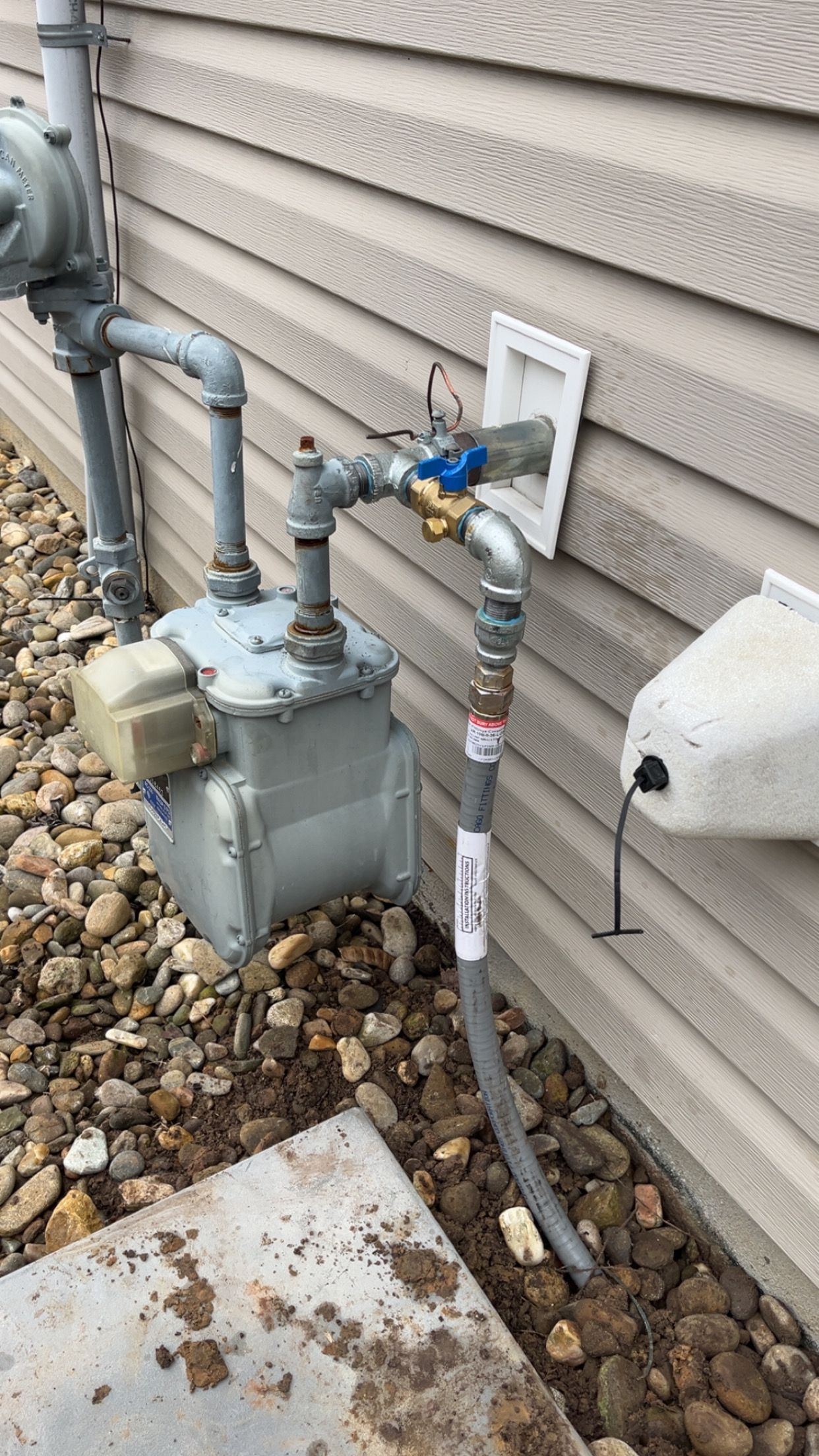 A gas meter with a connected yellow-handled shut-off valve and piping against a tan vinyl-sided wall over rocky ground.