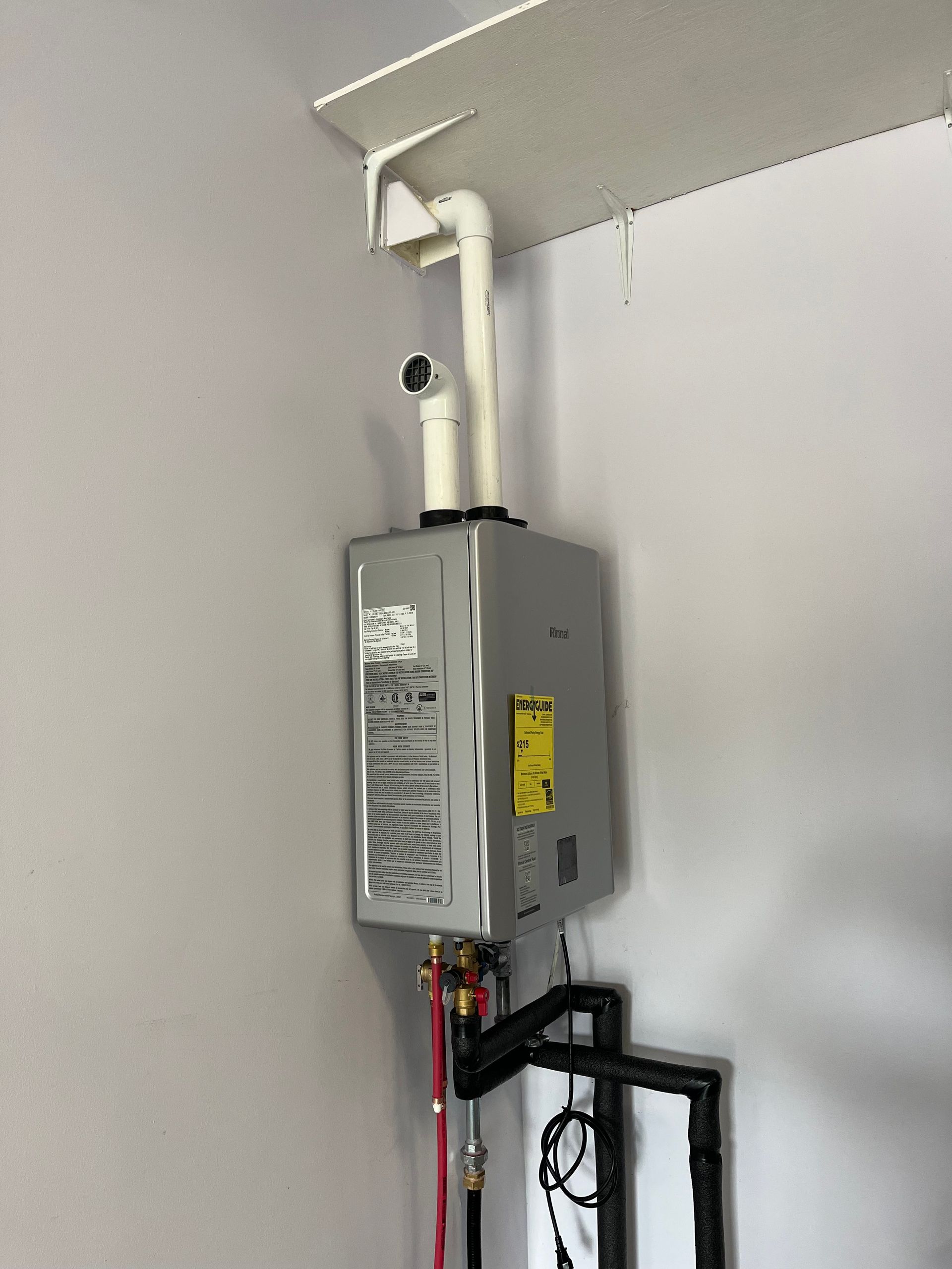 A tankless water heater mounted on an interior wall with connected white PVC venting and black plumbing pipes below.