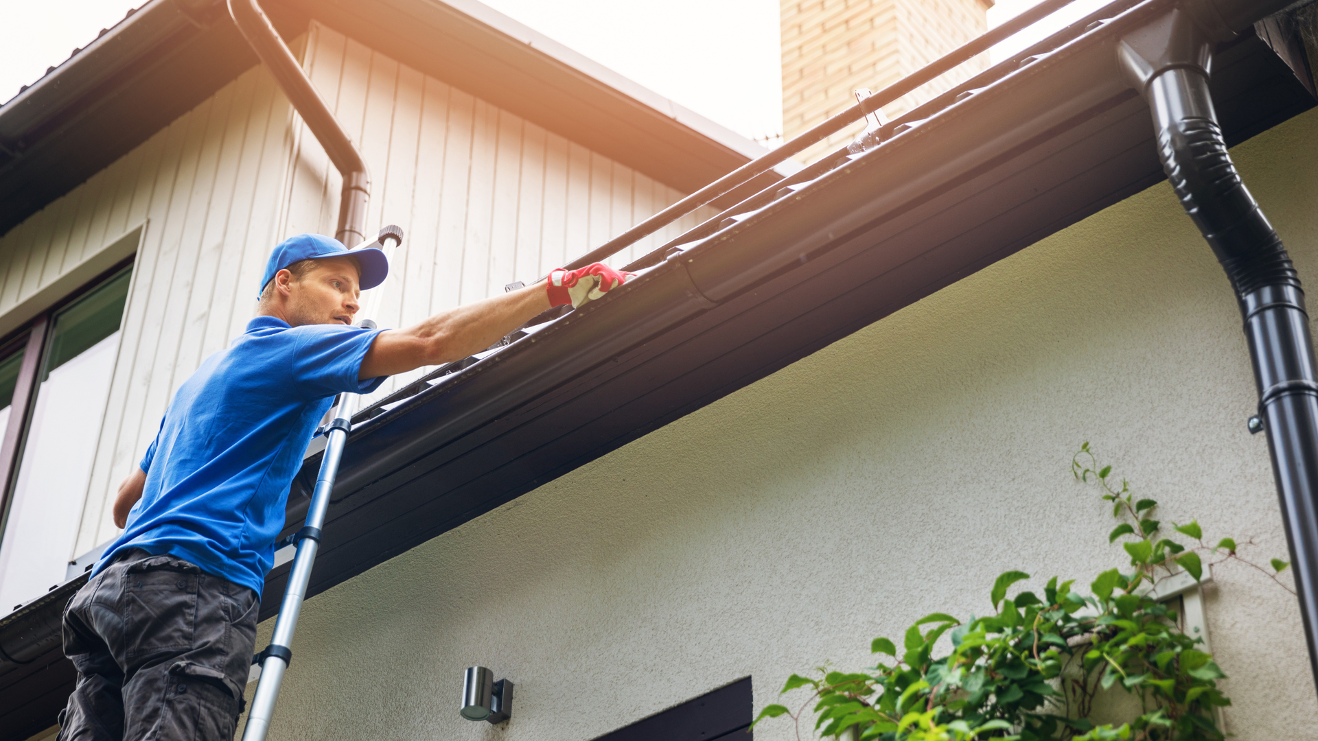 Technician cleaning house gutters from a ladder to prevent overflow and leaks.