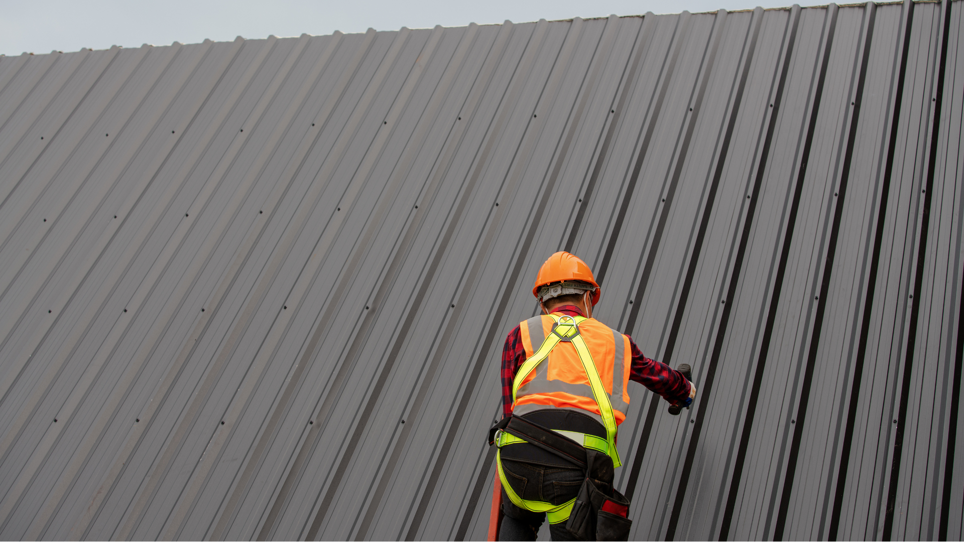 Worker in orange safety gear climbing a steep gray metal roof with a harness