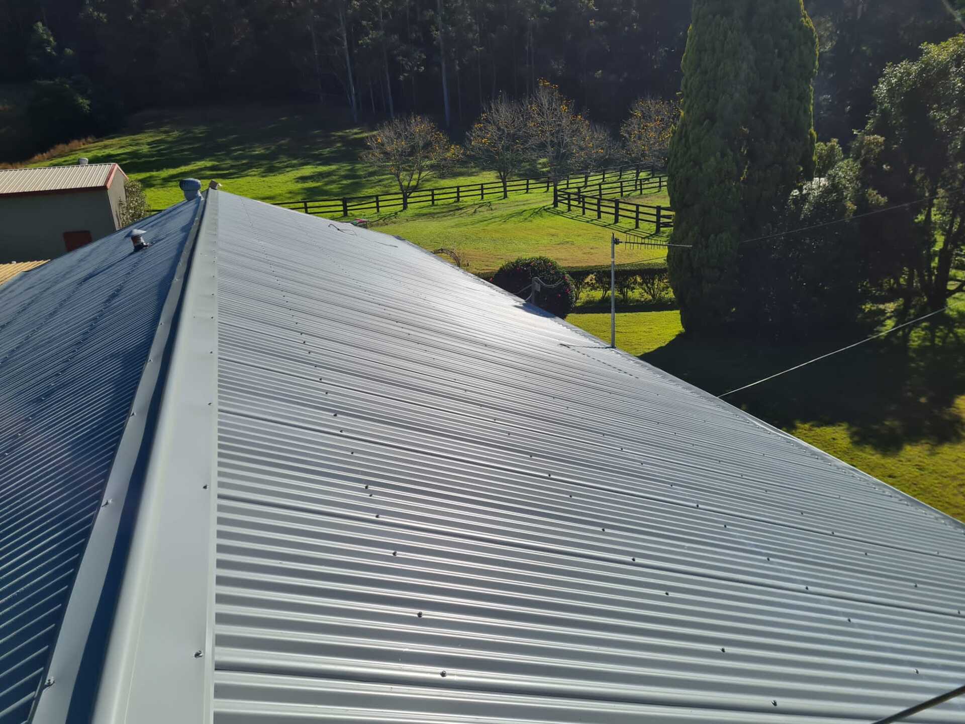 A top-down view of a grey corrugated metal roof on a sunny day, overlooking a rural landscape with trees and grass.