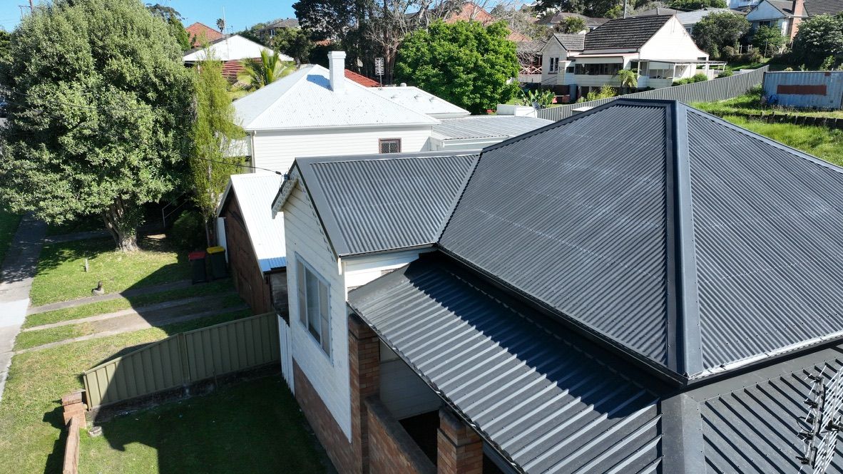 A high-angle view of a white house with a dark, corrugated metal roof in a residential neighborhood.