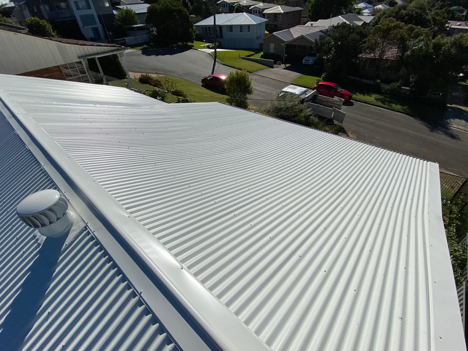 A high-angle view of a white, corrugated metal roof with a ventilation cap, overlooking a suburban street with cars.