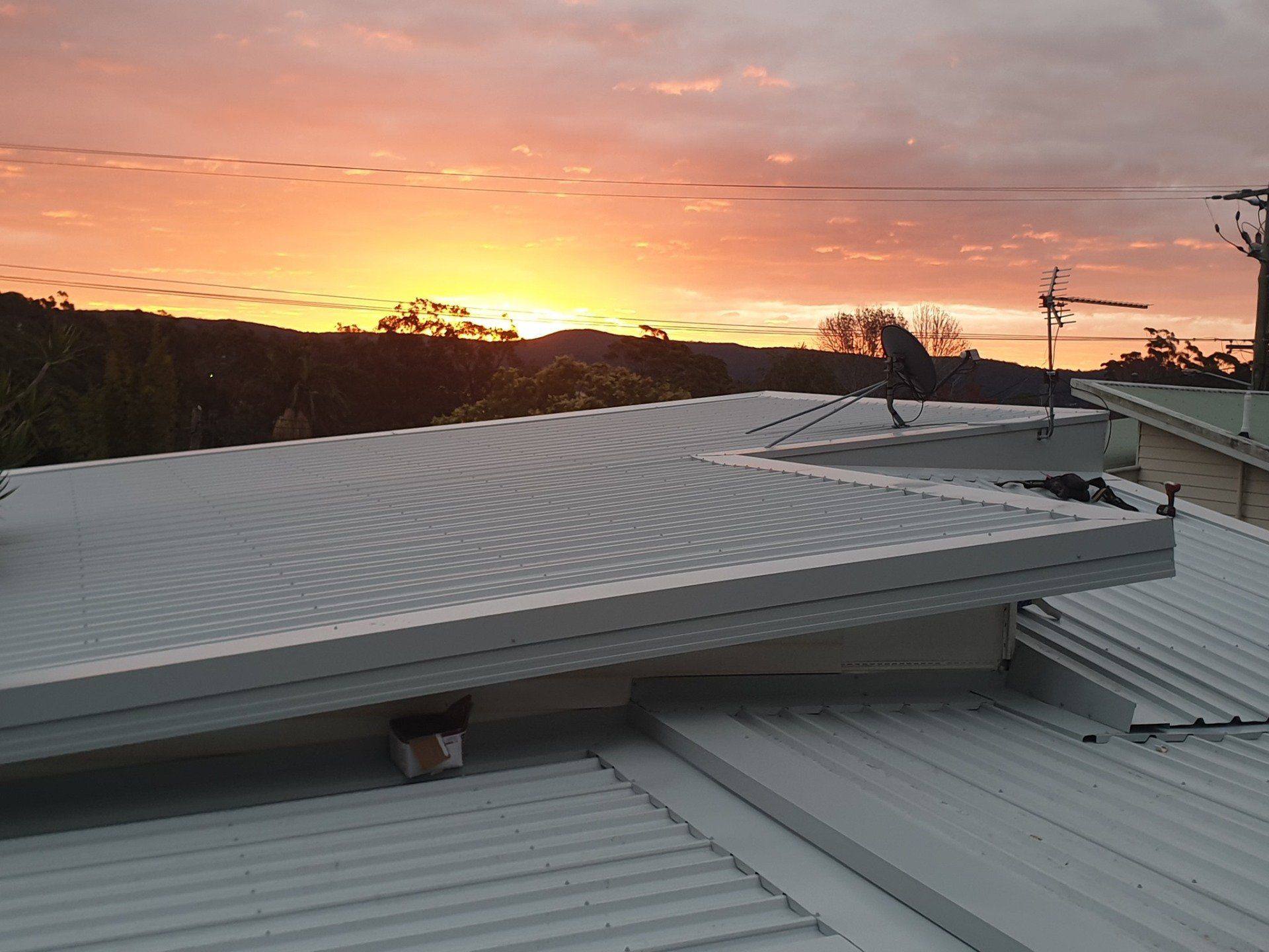 A sunrise with orange and pink clouds visible over a residential metal roof and antenna.