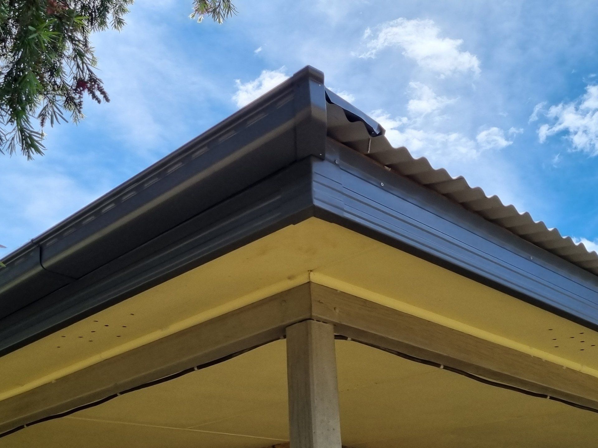 Low angle view of a house roof corner with dark grey corrugated metal roofing, matching gutters, and a beige soffit.