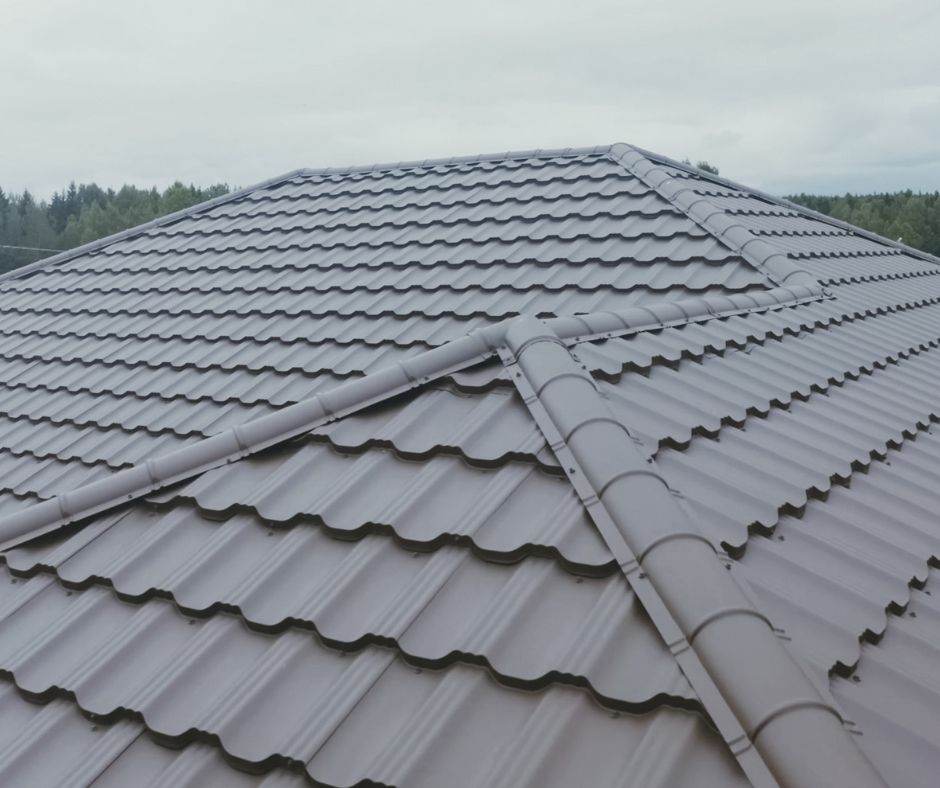 A close-up, high-angle view of a grey metal roof with a tiled pattern, showing hip rafters against a cloudy sky.