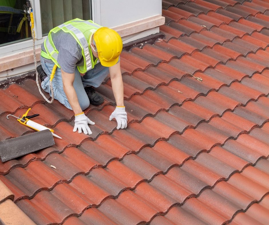 A construction worker in a hard hat and safety vest repairs terracotta roof tiles with sealant.