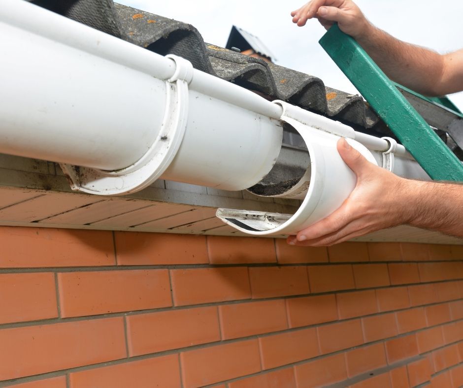 Hands repairing a broken white gutter section on a brick house exterior.