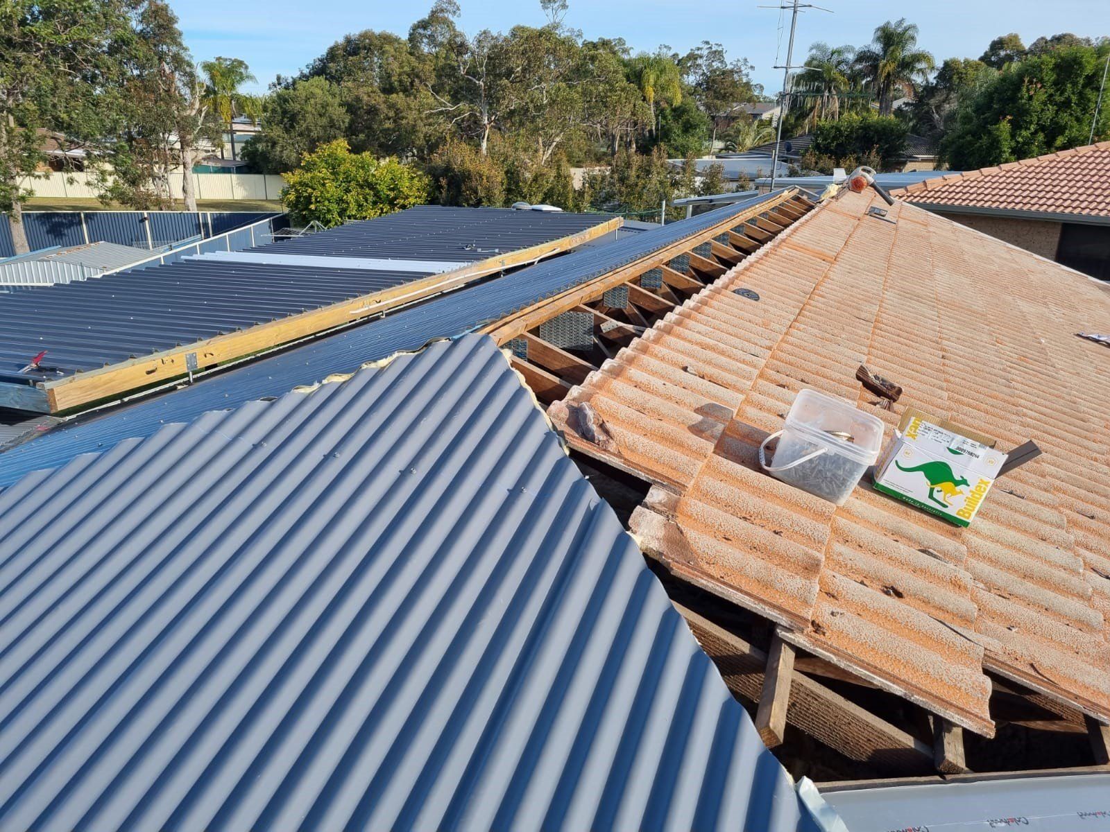 A residential roof under repair, showing a mix of new corrugated grey metal sheeting and old terracotta roof tiles.