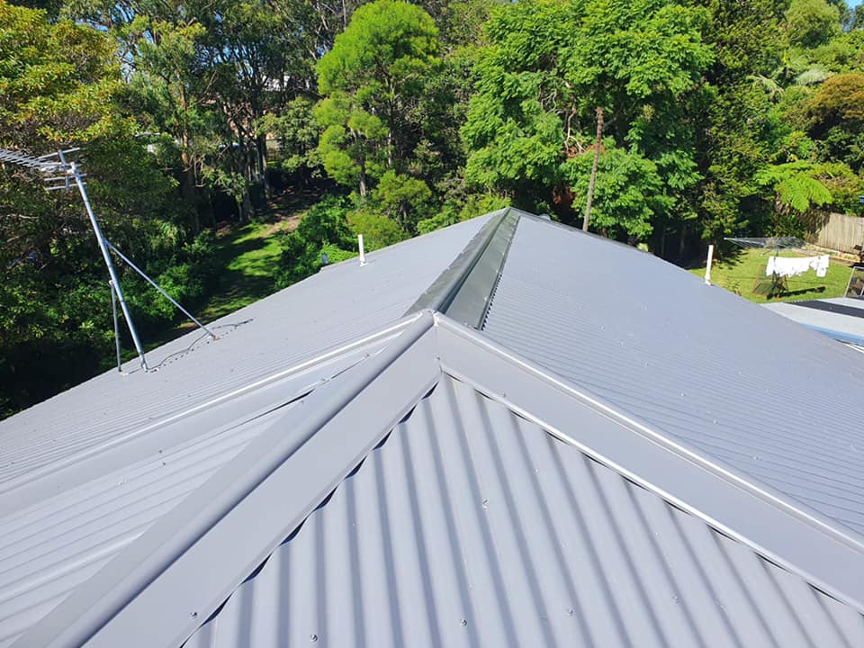 A view from a roof showing grey corrugated metal sheets, a central ridge cap, and surrounding green trees.
