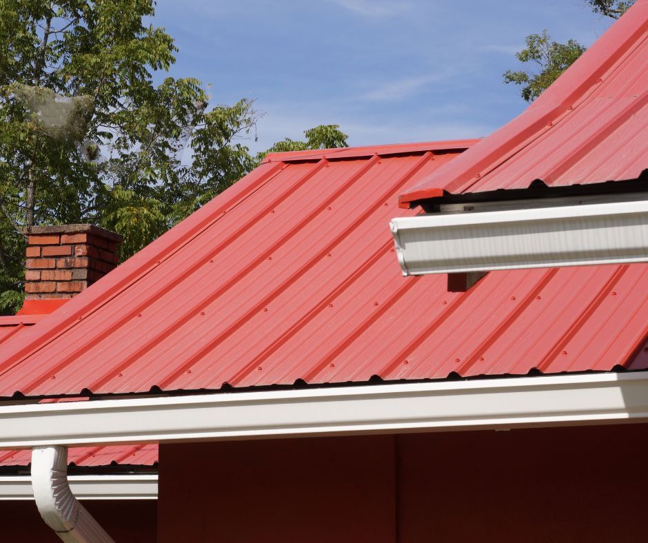 A bright red metal roof on a house with a white gutter system against a blue sky and green trees.