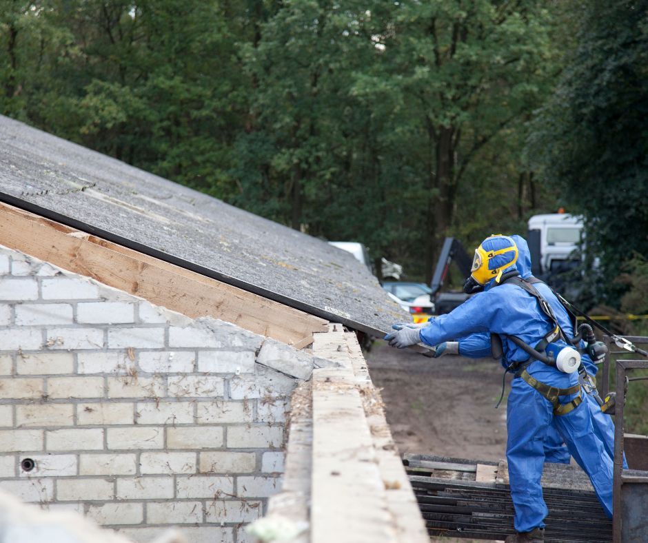 A worker in protective blue coveralls and a respirator removes roofing panels from a building structure.