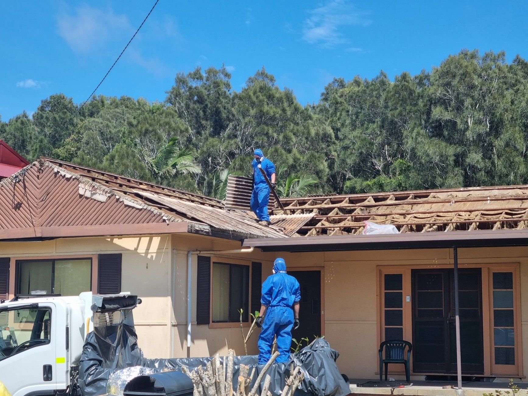 Two people in blue protective hazmat suits working on a house roof that is being stripped and repaired.