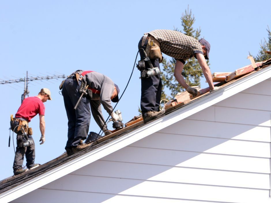 Three roofers work on a pitched house roof under a clear blue sky, using pneumatic tools to install shingles.