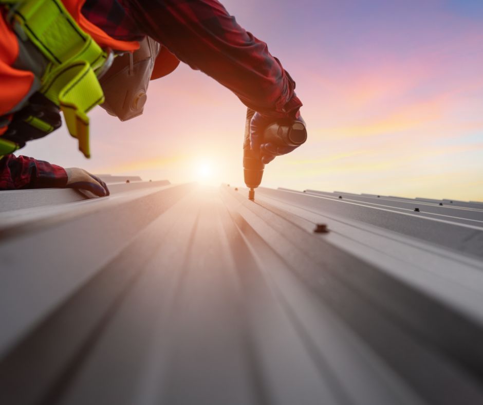 A construction worker in a high-visibility vest uses a drill to secure metal roofing panels against a sunset backdrop.