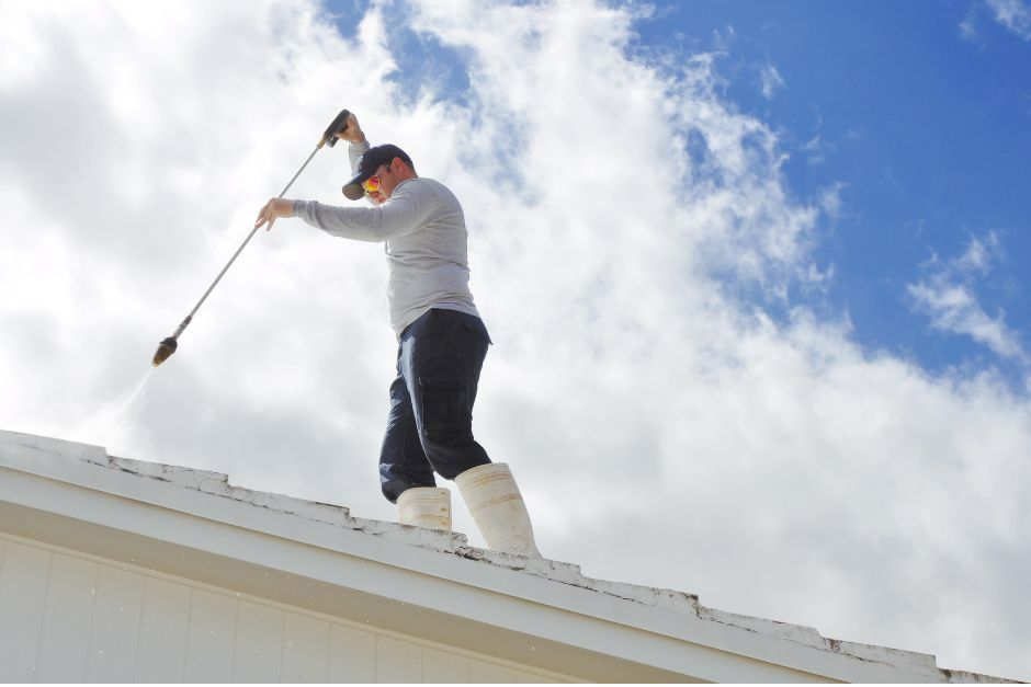 A person standing on a sloped roof uses a pressure washer to clean the surface against a bright, cloudy sky.