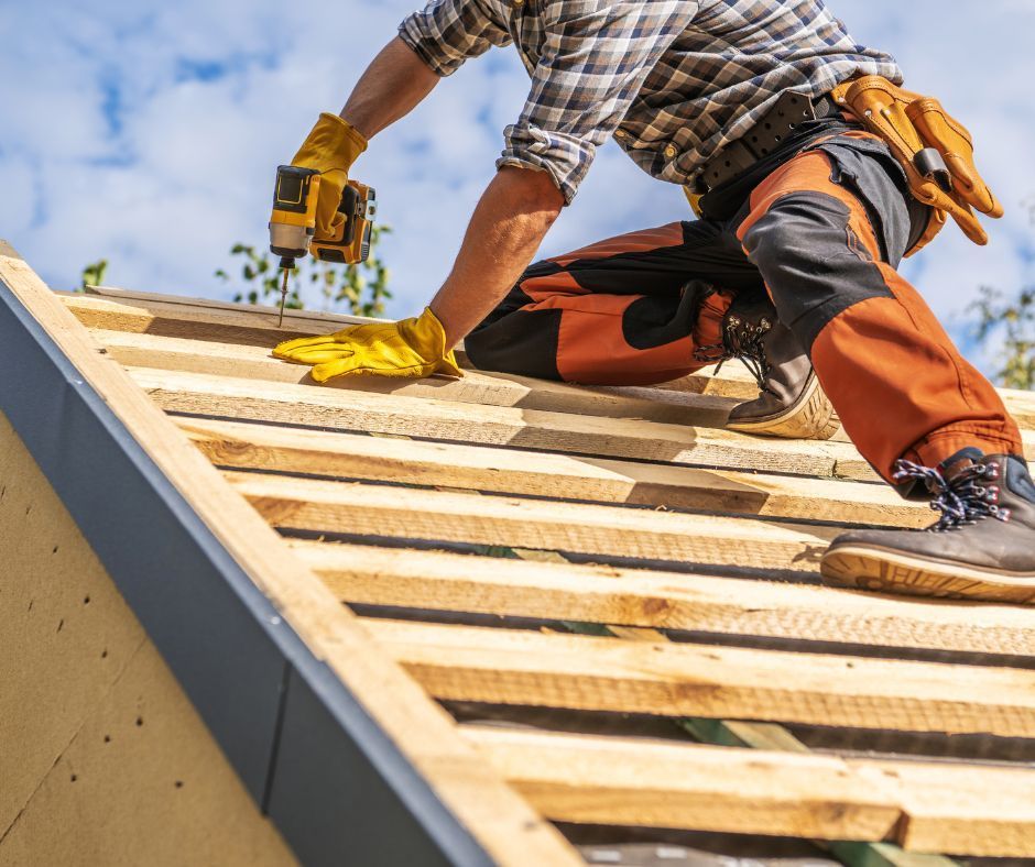 A person in work clothes and yellow gloves using a power drill on a wooden roof frame against a blue sky.