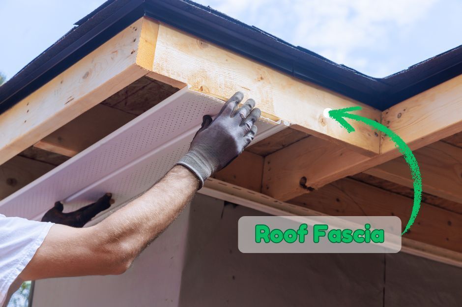 A worker installing white soffit material under the roof eaves, with an arrow pointing to the wooden roof fascia.