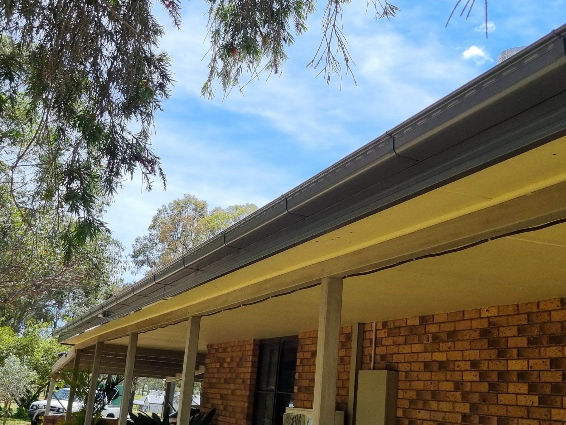 A dark brown gutter running along the edge of a yellow-painted roof eave above a brick wall and outdoor porch.