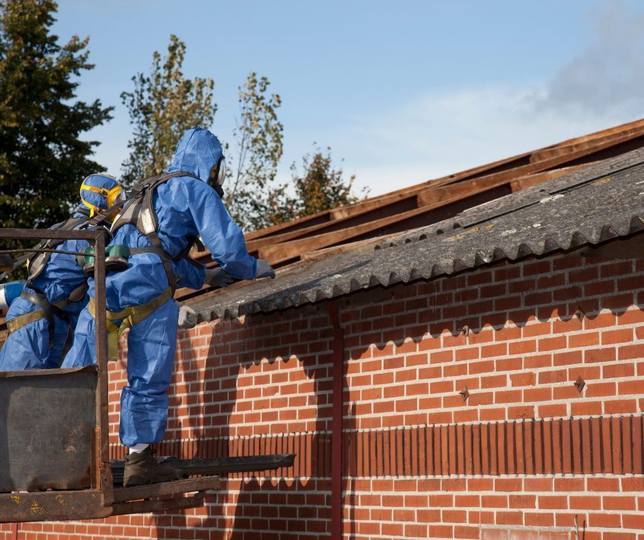 Two workers in blue protective suits and respirators remove corrugated roofing material from a brick building.