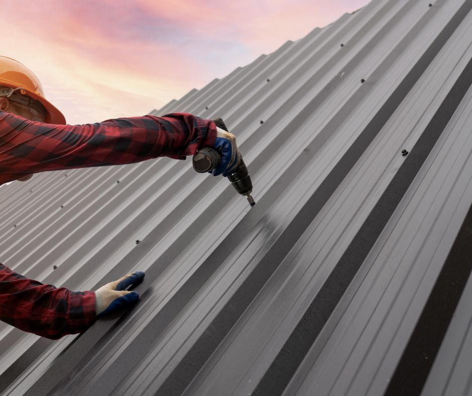 A worker in a red plaid shirt and safety helmet uses a drill to secure metal roofing panels at sunset.