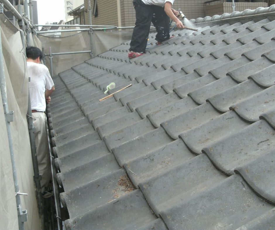 Two workers on a scaffolding-enclosed house roof, one using a pressure washer on grey clay tiles.